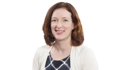 A person with shoulder-length brown hair, wearing a white cardigan over a patterned top, poses against a plain white background. They are smiling and looking at the camera.