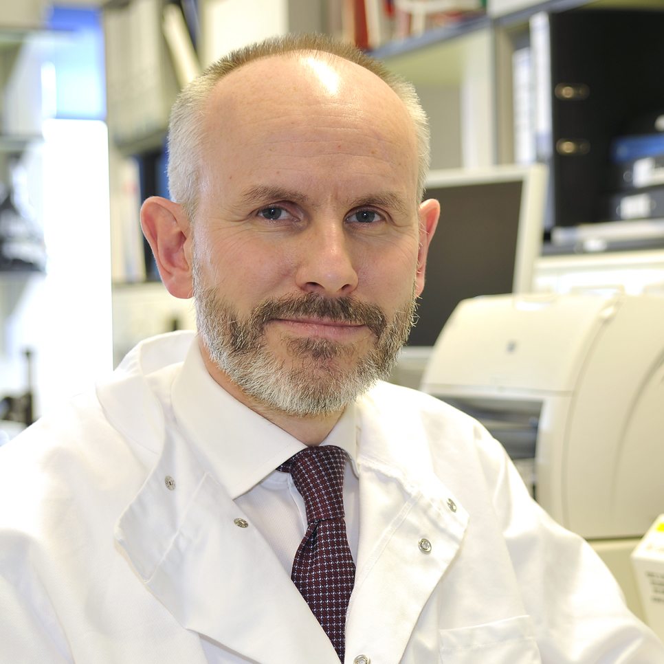 A man with a beard and short hair, wearing a white lab coat embossed with the NA-ATTC logo and a tie, is seated in a laboratory setting. Shelves filled with books and equipment are visible in the background.