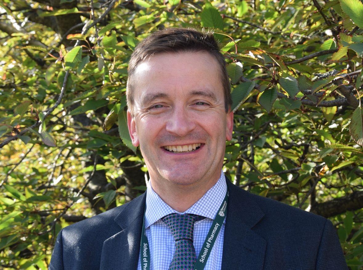 A person with short brown hair is smiling while standing outdoors in front of a tree. They are wearing a dark suit, blue checkered shirt, and a green tie. The NA-ATTC lanyard around their neck adds a touch of distinction against the backdrop of vibrant green leaves.