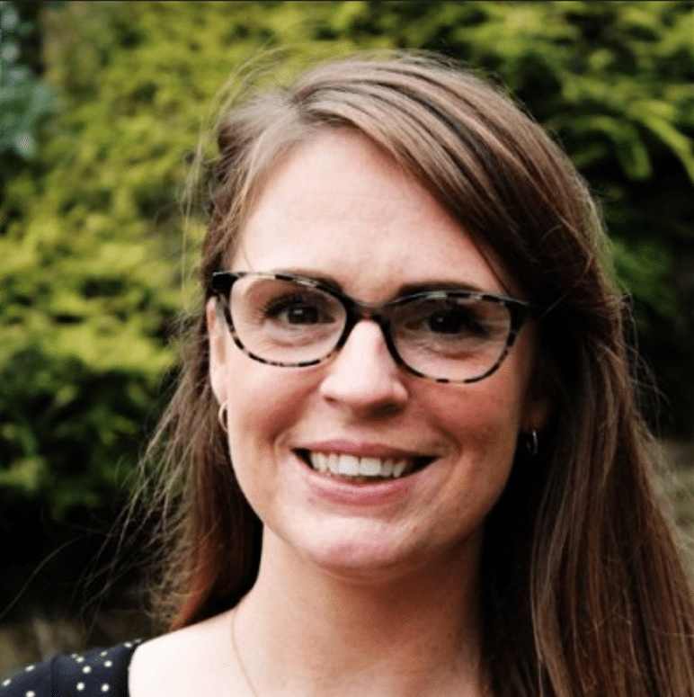 A woman with long brown hair and glasses smiles at the camera. She is wearing a black top with white polka dots. The background is lush with greenery.