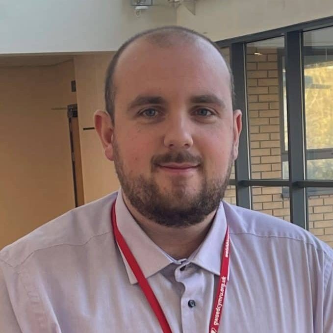 A person with a beard and short hair is wearing a light-colored shirt and a red lanyard, standing indoors with a blurred background of a window and beige walls.