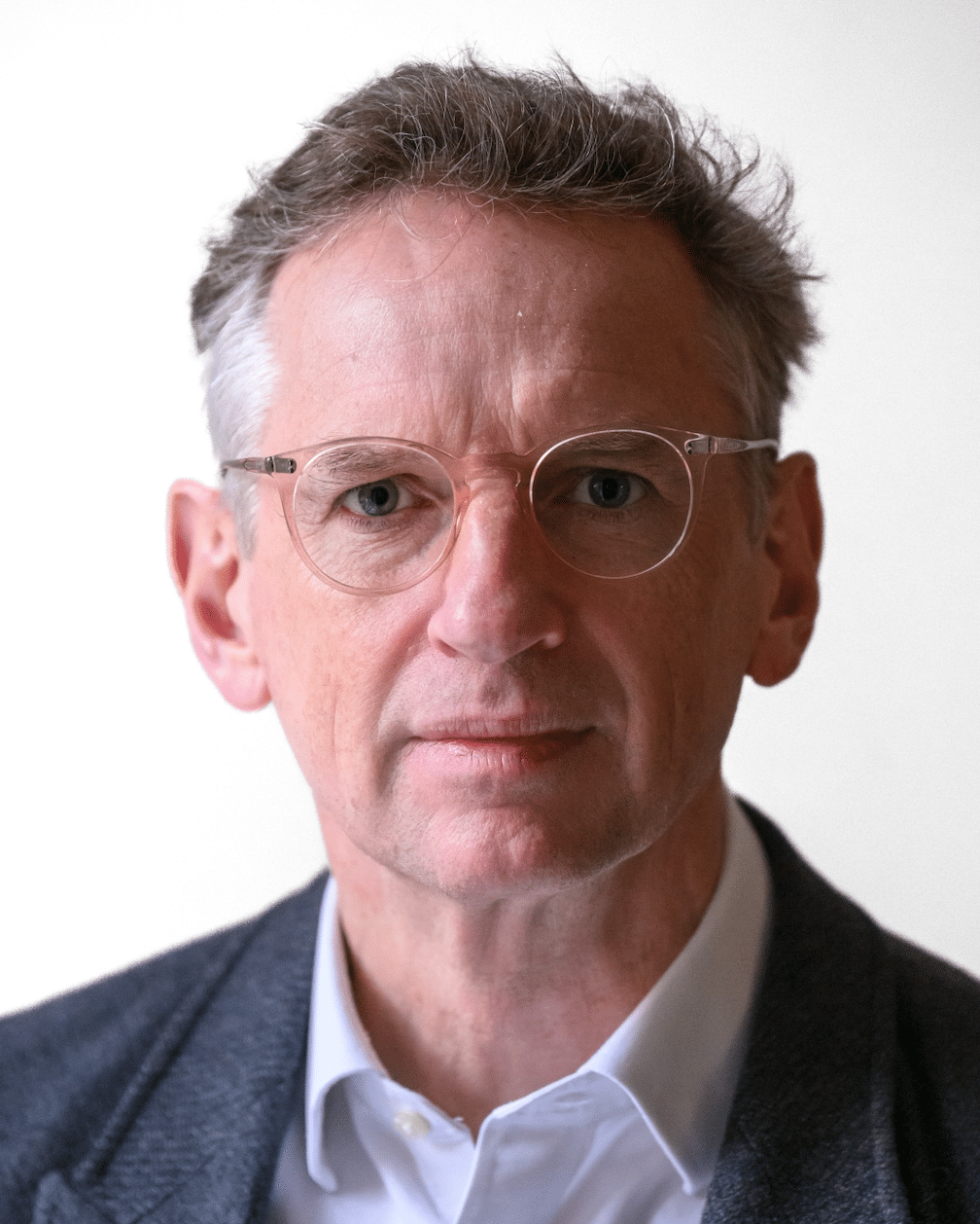A person with short hair and glasses is wearing a dark blazer over a white shirt. They are looking directly at the camera with a neutral expression, set against a plain background.