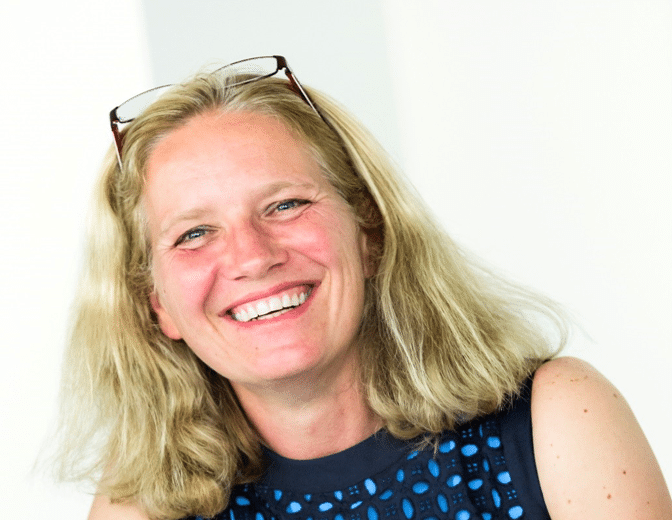 Smiling woman with shoulder-length blonde hair, wearing glasses on her head and a sleeveless blue dress with a patterned design. She is looking towards the camera, against a light background.