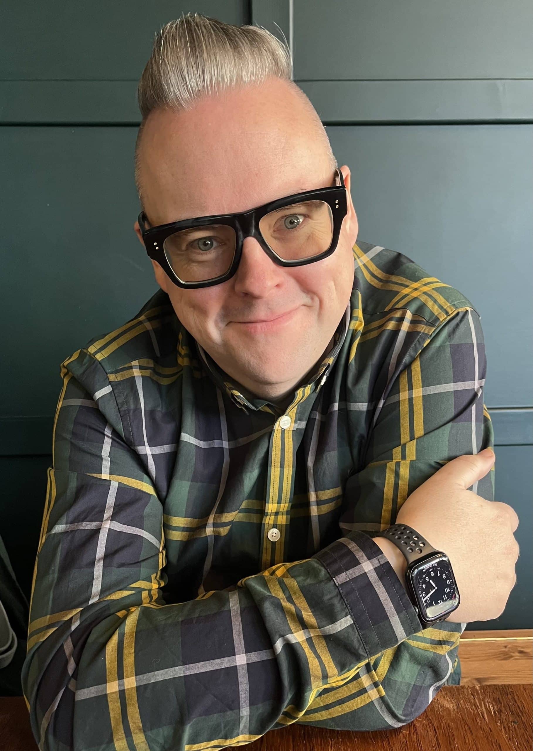 A person with short, styled hair and large black glasses is smiling while sitting at a table. They are wearing a green and yellow plaid shirt and a smartwatch. The background features a dark green wall.